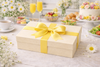 Beige gift box with a yellow ribbon on a table with flowers and fruit in the background